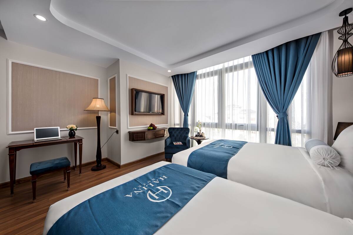 A hotel room with two beds, a desk with a laptop, a chair, a TV, and a window with curtains.