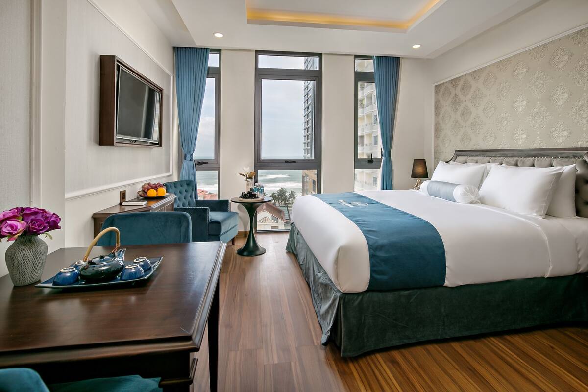 A hotel room with a large bed, a desk with a vase of flowers, a small table with a teapot, and a view of the city through large windows.
