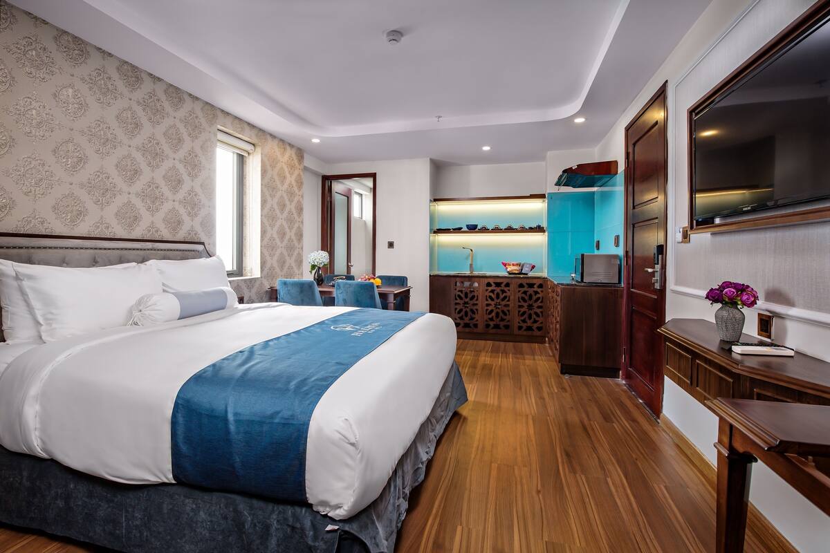 A modern hotel room with a large bed, a TV, a small kitchen area, and a bathroom.