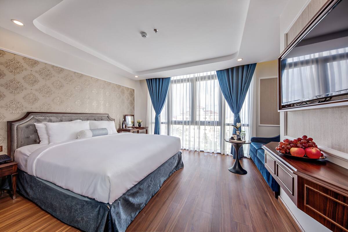 A hotel room with a large bed, a TV, a small table with a fruit bowl, and a view of the city through large windows.