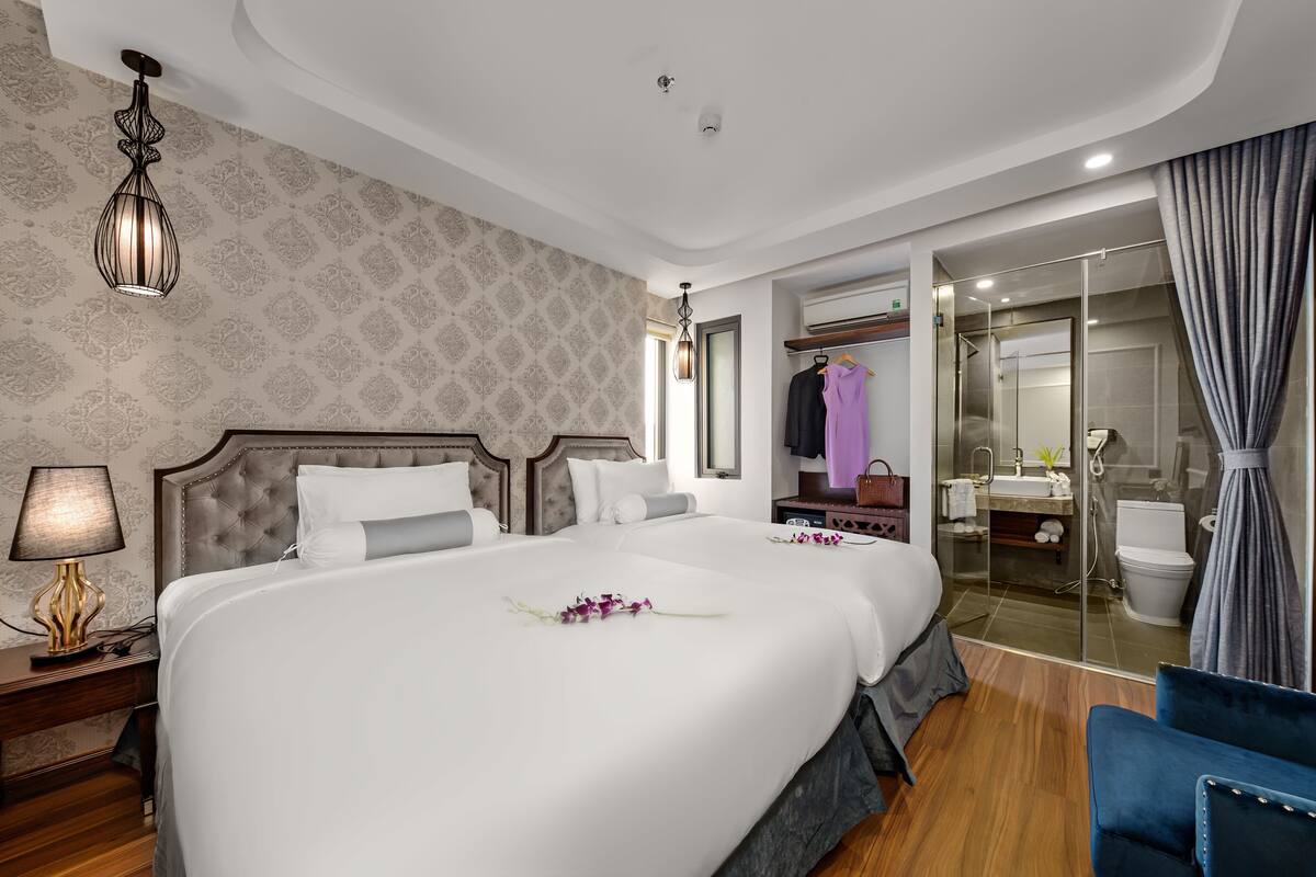 A hotel room with a large bed, a bedside table with a lamp, a wardrobe, a bathroom with a glass shower, and a blue chair.