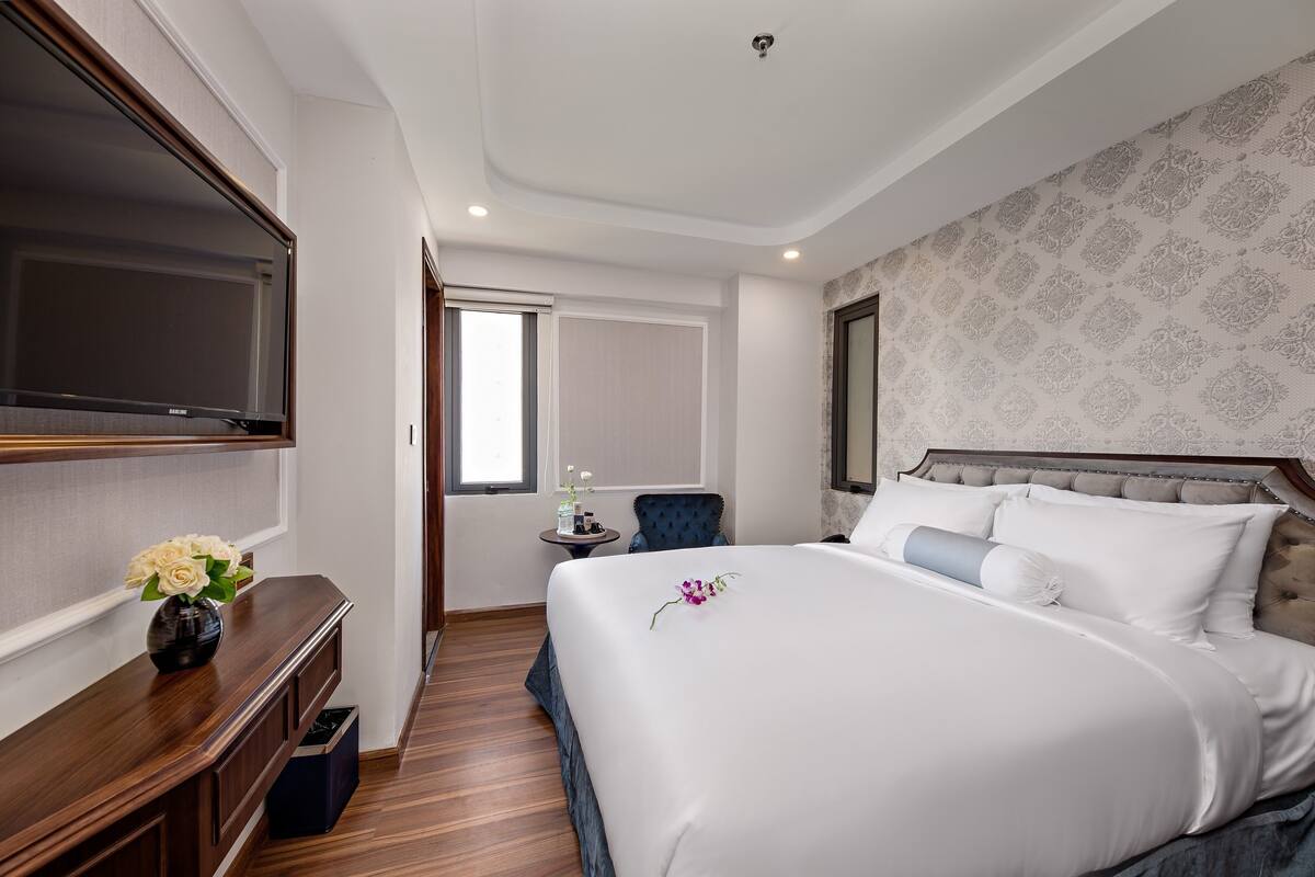 A hotel room with a large bed, a TV mounted on the wall, a small table with a vase of flowers, and a window with a view.