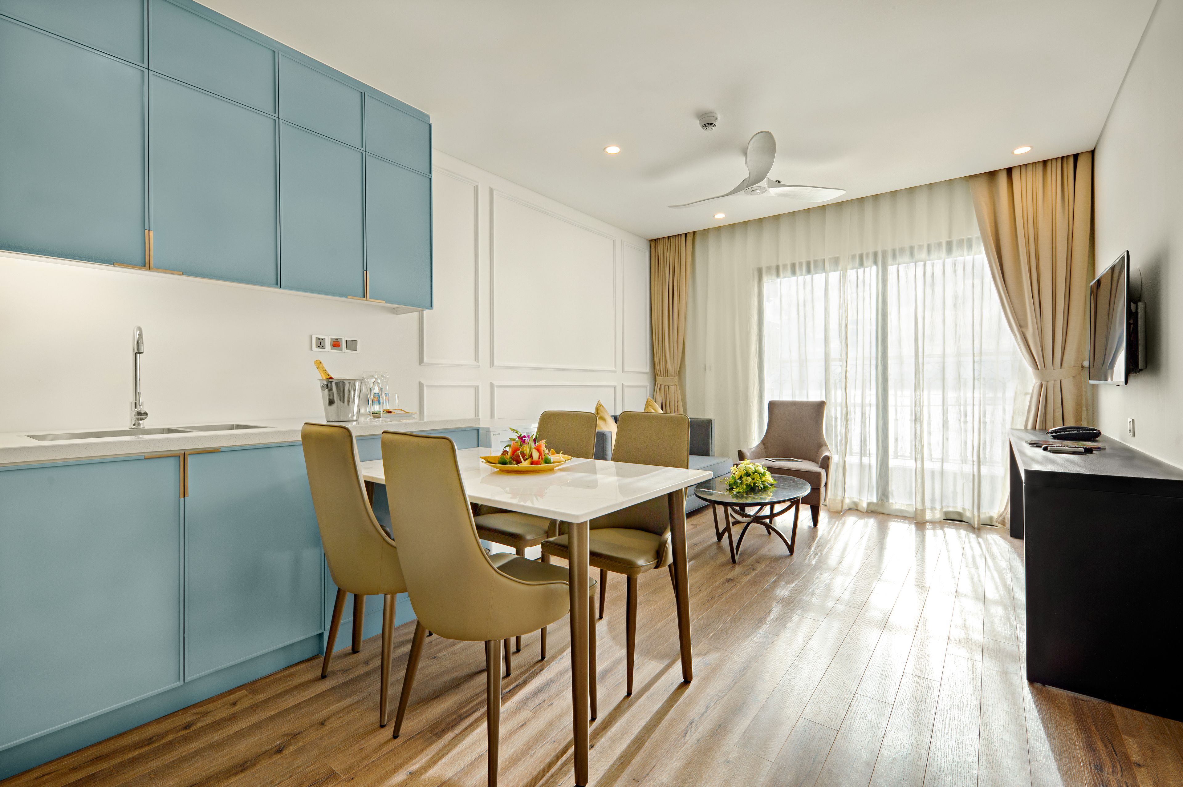 A modern kitchen with blue cabinets, a dining area with a white table and chairs, and a living area with a sofa and a TV.