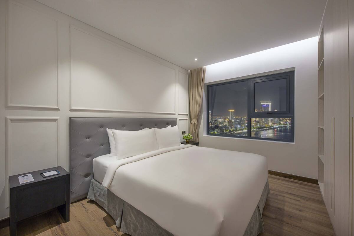 Quarto de hotel bem arrumado com uma cama grande, mesa de cabeceira, janela com vista para a cidade e design moderno.