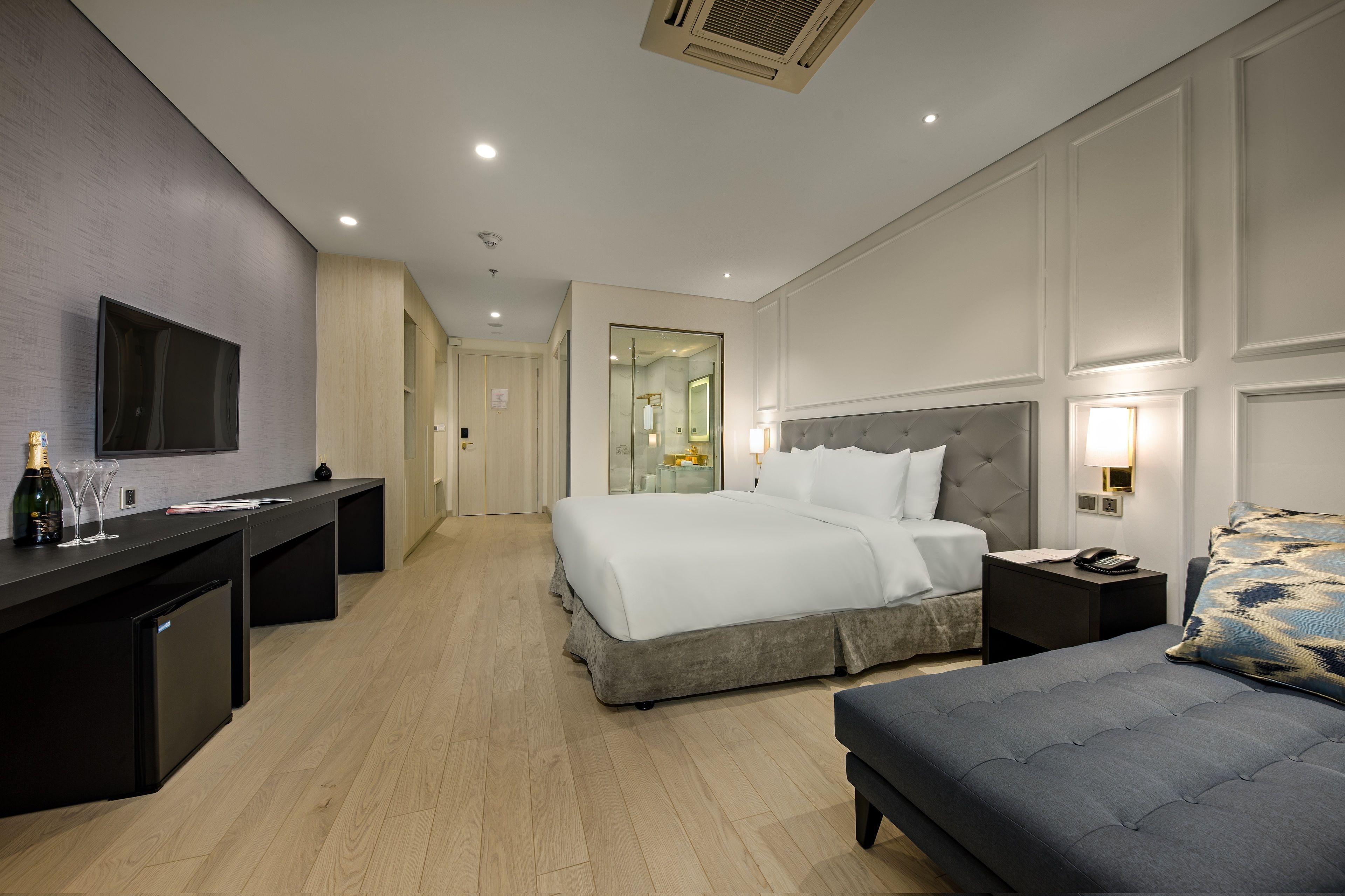 A modern hotel room with a large bed, bedside tables, a desk with a flat-screen TV, and a minibar.