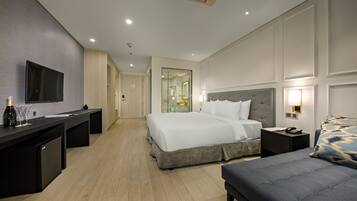 A modern hotel room with a large bed, bedside tables, a desk with a flat-screen TV, and a minibar.