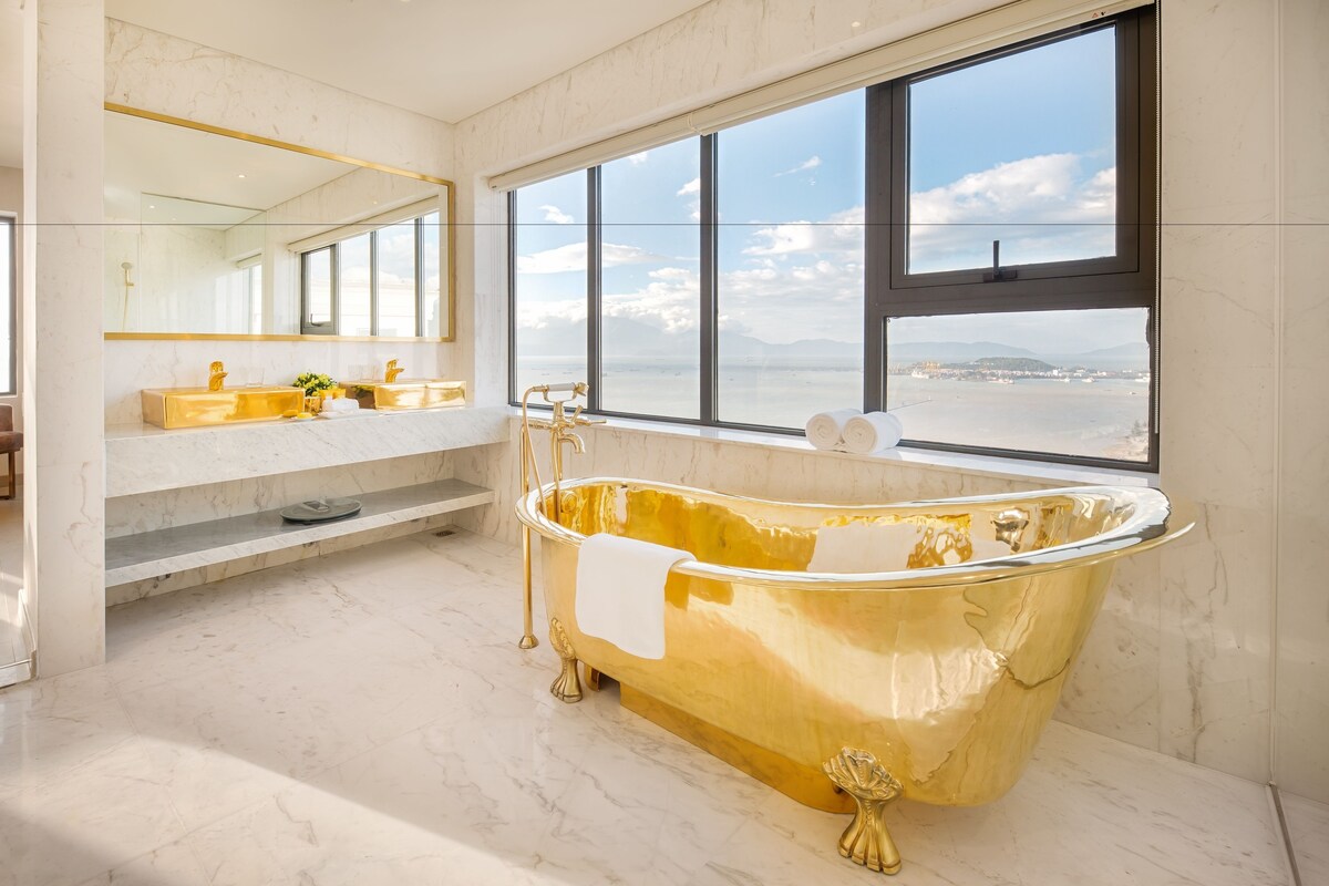 A luxurious bathroom with a gold bathtub, large windows, and a marble floor.