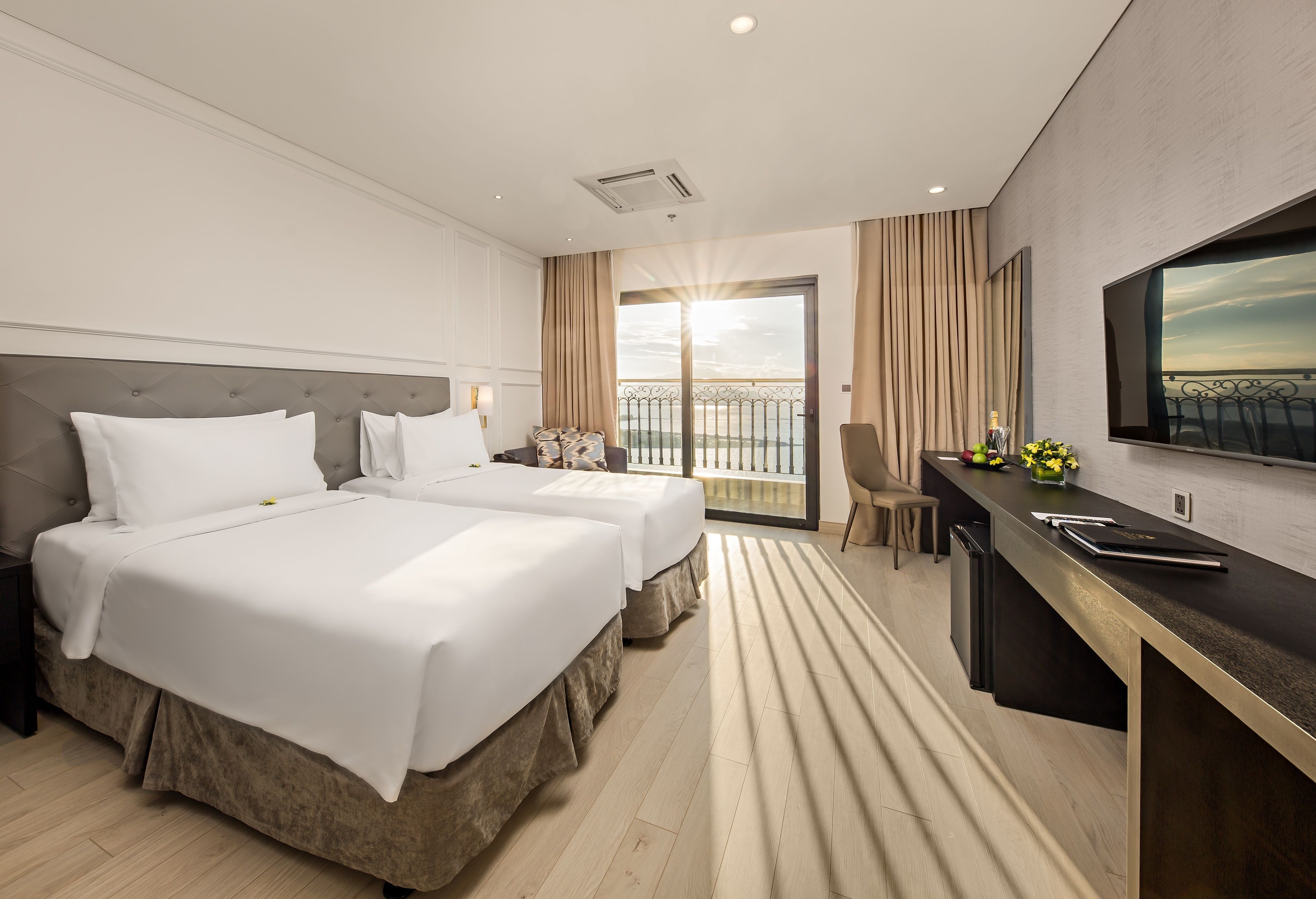 A hotel room with two beds, a balcony with a view, a desk with a chair, and a TV mounted on the wall.