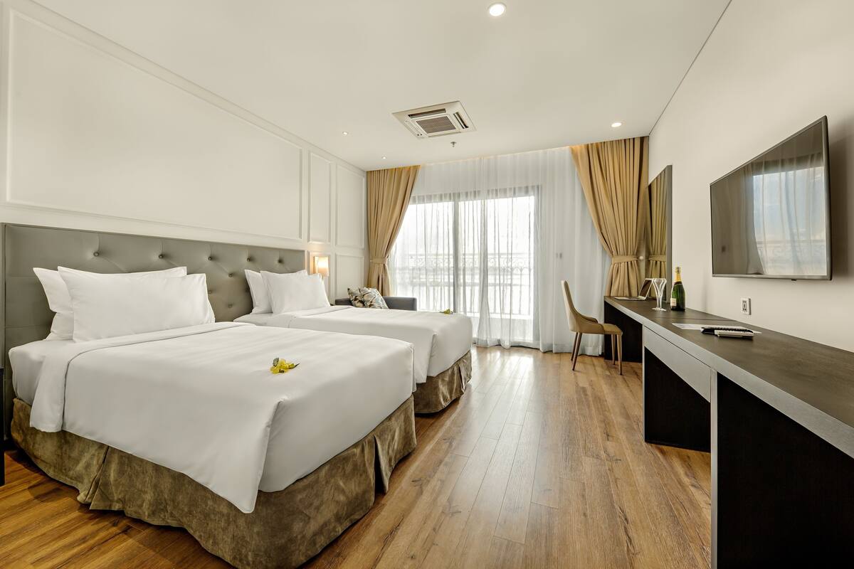 Quarto de hotel com duas camas, uma escrivaninha, uma cadeira, televisão e uma janela grande com cortinas.