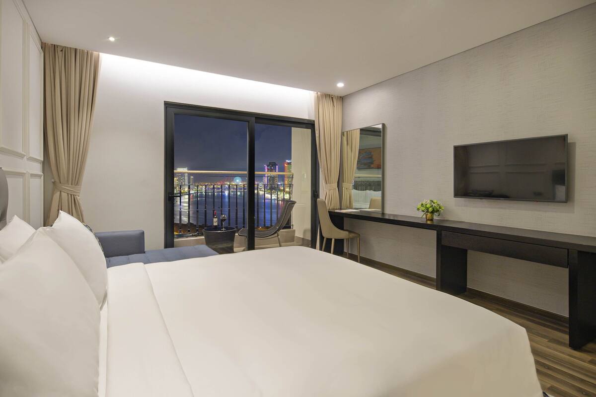 Quarto de hotel com uma cama grande, uma escrivaninha, uma cadeira, uma televisão e uma varanda com vista para a cidade.