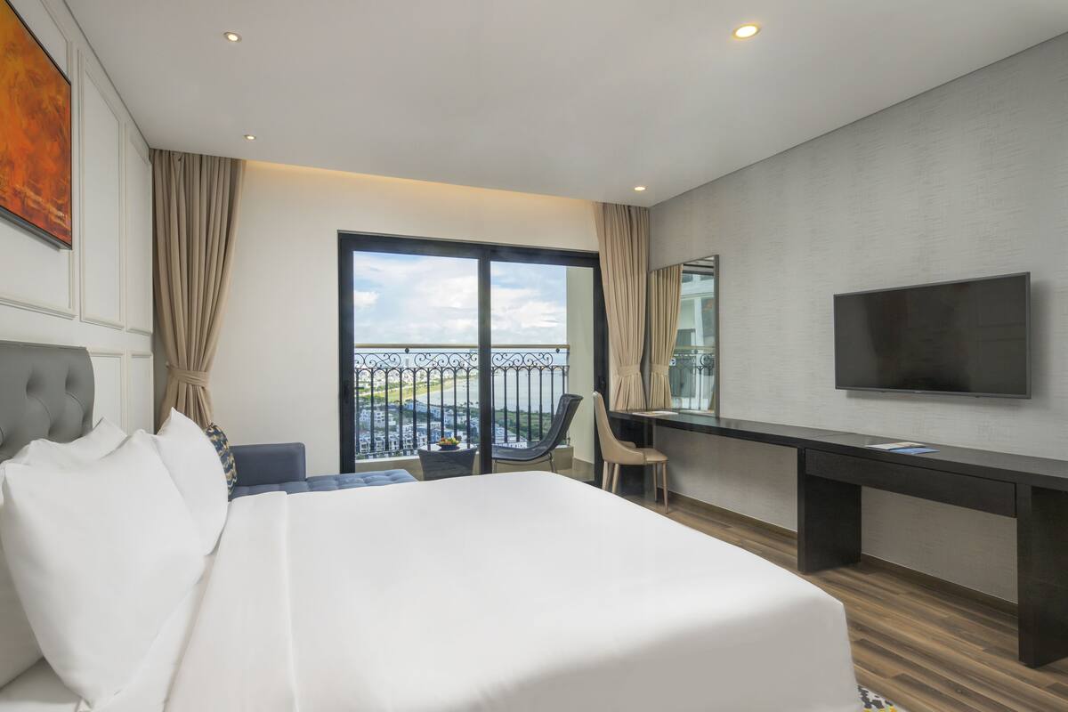 Quarto de hotel com cama grande, escrivaninha, cadeira, televisão e varanda com vista para a cidade.
