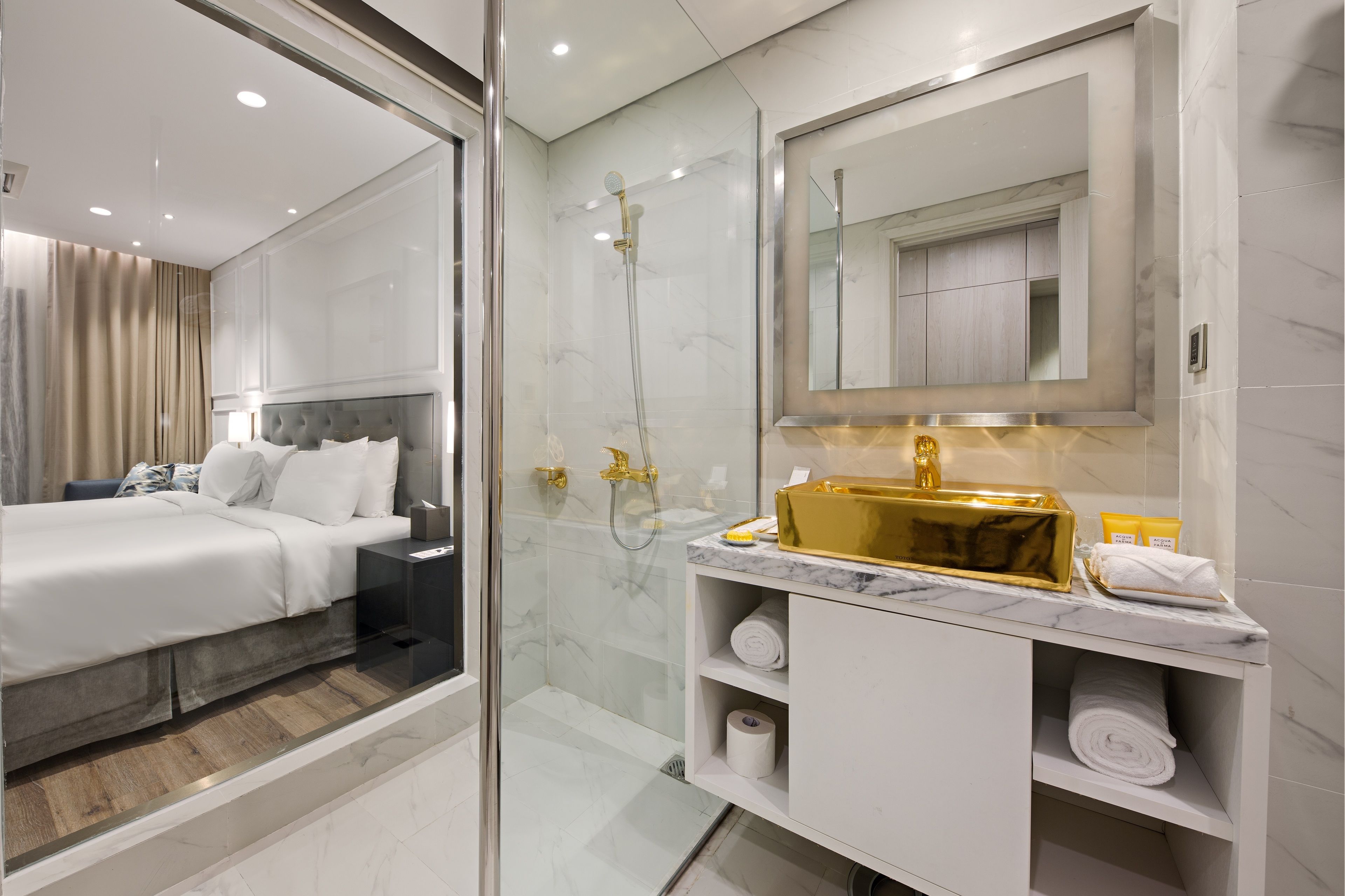 Une chambre d’hôtel moderne dotée d’une douche en verre, d’un grand lit avec du linge de lit blanc, d’un meuble-lavabo en marbre dans la salle de bain et d’un grand miroir au-dessus du lavabo.