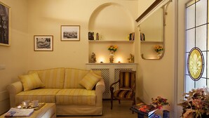Een gezellige woonkamer met een gestreept bankje, een houten stoel en een glazen salontafel. Er hangen ingelijste foto's aan de muur, een boekenkast en een spiegel.