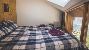 Une chambre avec un grand lit, une lucarne, une fenêtre donnant sur un beau paysage et une porte en bois.