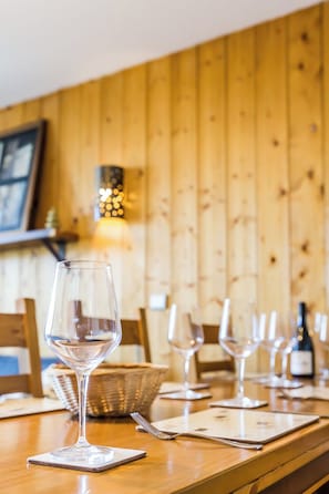 A wooden dining table with wine glasses and a bottle, set against a wooden wall with a wall-mounted light fixture.