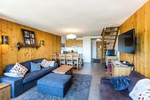 Ett modernt vardagsrum med en soffa, en pall och ett soffbord, träpanel på väggarna, en matplats och en tv som är monterad på väggen.
