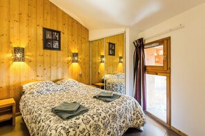 Habitación de hotel con pared de paneles de madera, una cama con colcha estampada, dos mesitas de noche, un espejo y una ventana con cortinas.