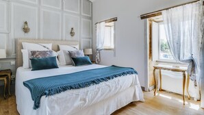 Een slaapkamer met een groot bed, een kroonluchter, een raam met gordijnen en een houten tafel.