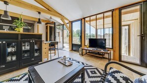 Een moderne woonkamer met een bank, salontafel en televisie. Er is een keukenhoek met kastjes en een eettafel. Grote ramen zorgen voor natuurlijk licht en uitzicht op de omgeving.