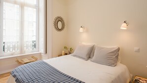 Une chambre avec un grand lit, une fenêtre avec des rideaux légers, un miroir fixé au mur, deux appliques murales et un luminaire au plafond.