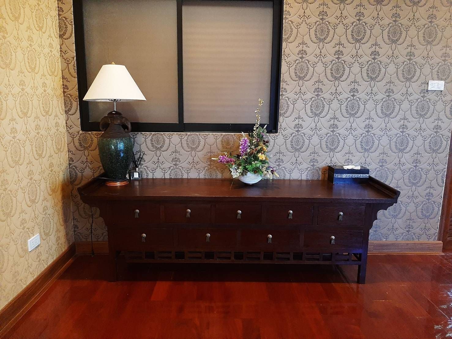 A wooden console table with a lamp, a vase, and a flower arrangement against a patterned wallpaper.