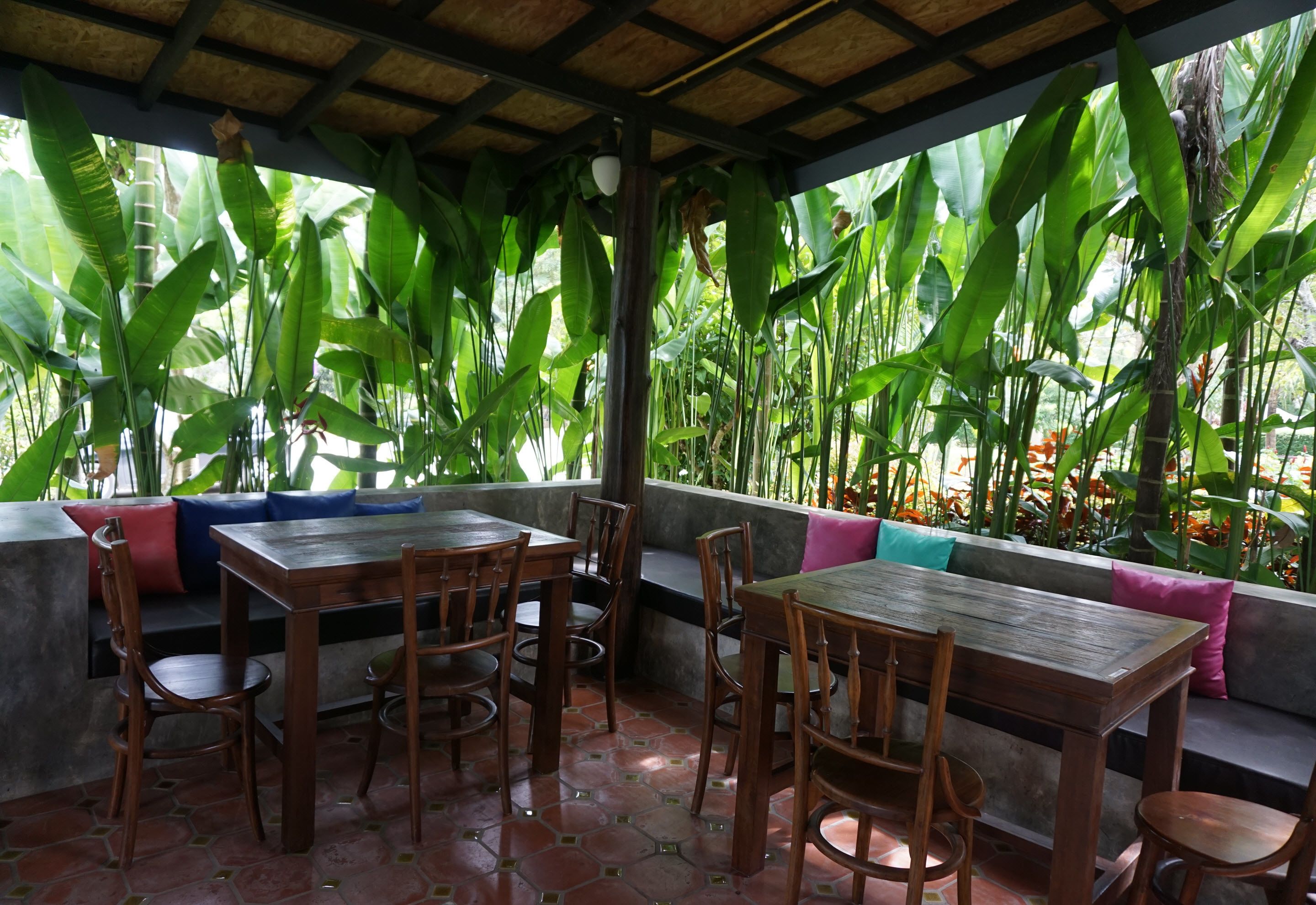 A covered outdoor seating area with wooden tables and chairs, surrounded by lush green plants and a view of the outdoors.
