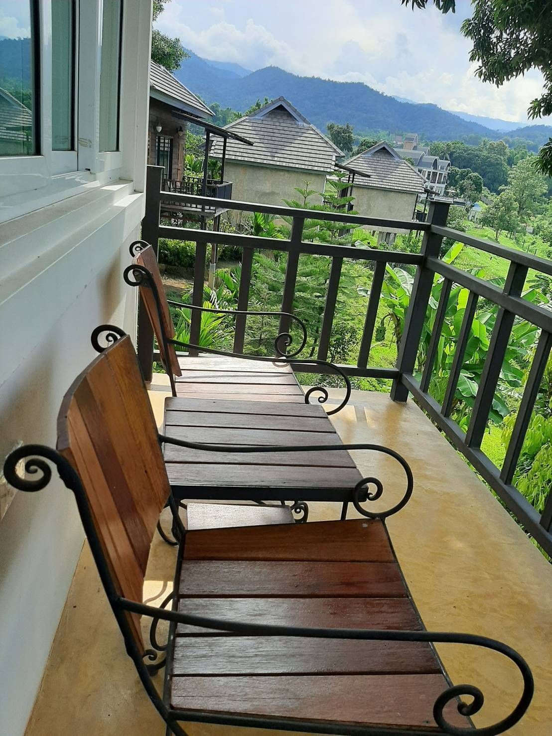 A wooden bench with metal armrests on a balcony with a view of lush greenery and mountains.