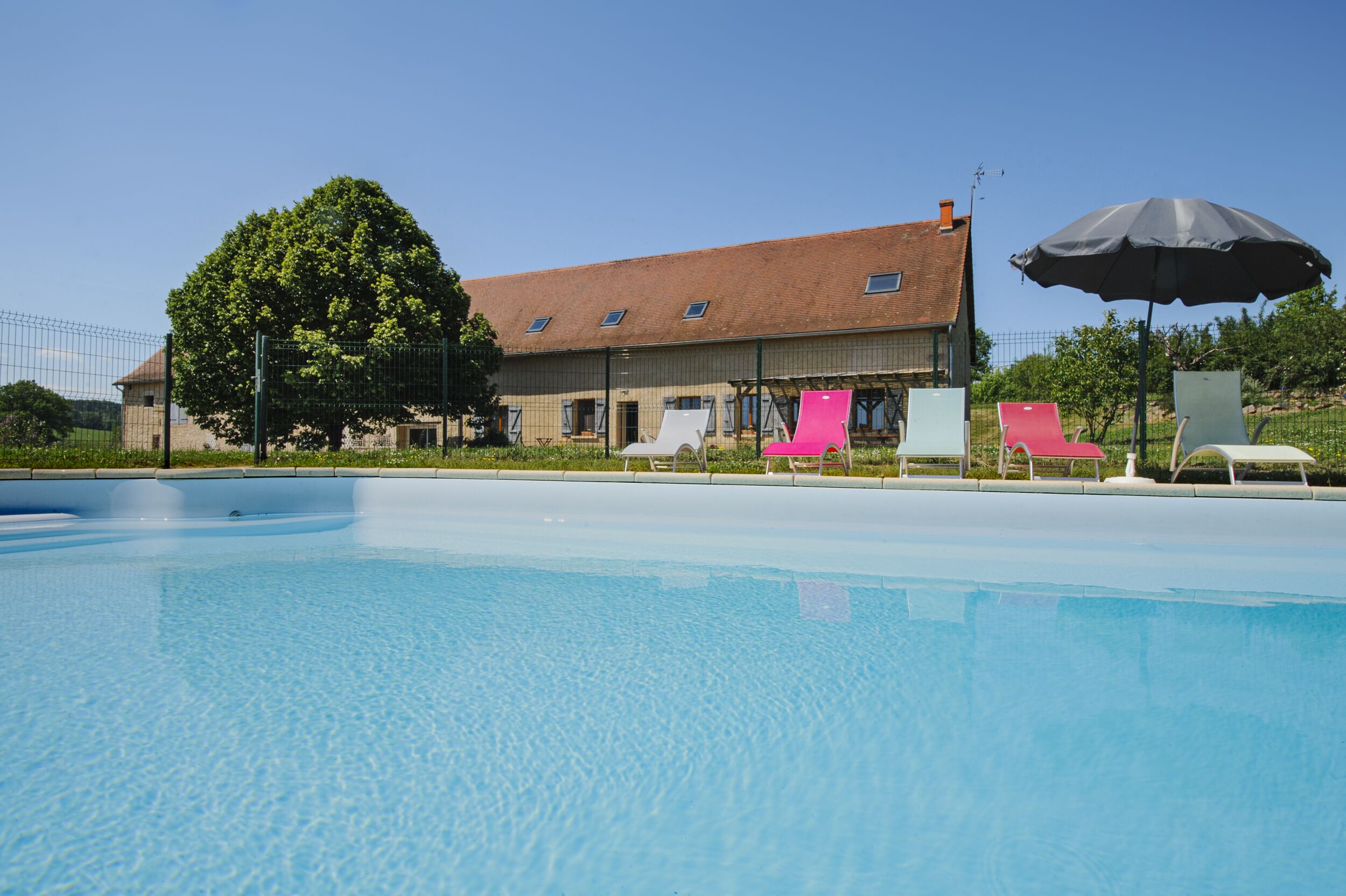 Una piscina con sdraio e un ombrellone, un albero grande e un edificio con tetto spiovente.