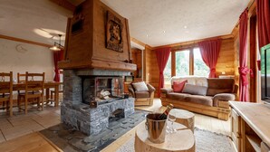 Een gezellig chaletinterieur met een stenen openhaard, houten meubels en grote ramen met rode gordijnen.