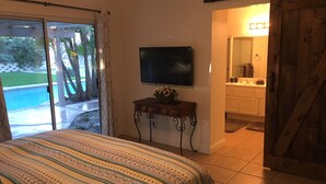 A bedroom with a bed, a TV, a small table with a vase, and a bathroom visible through an open door.