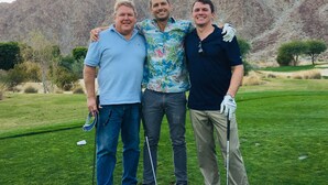 Three people standing on a golf course with mountains in the background.