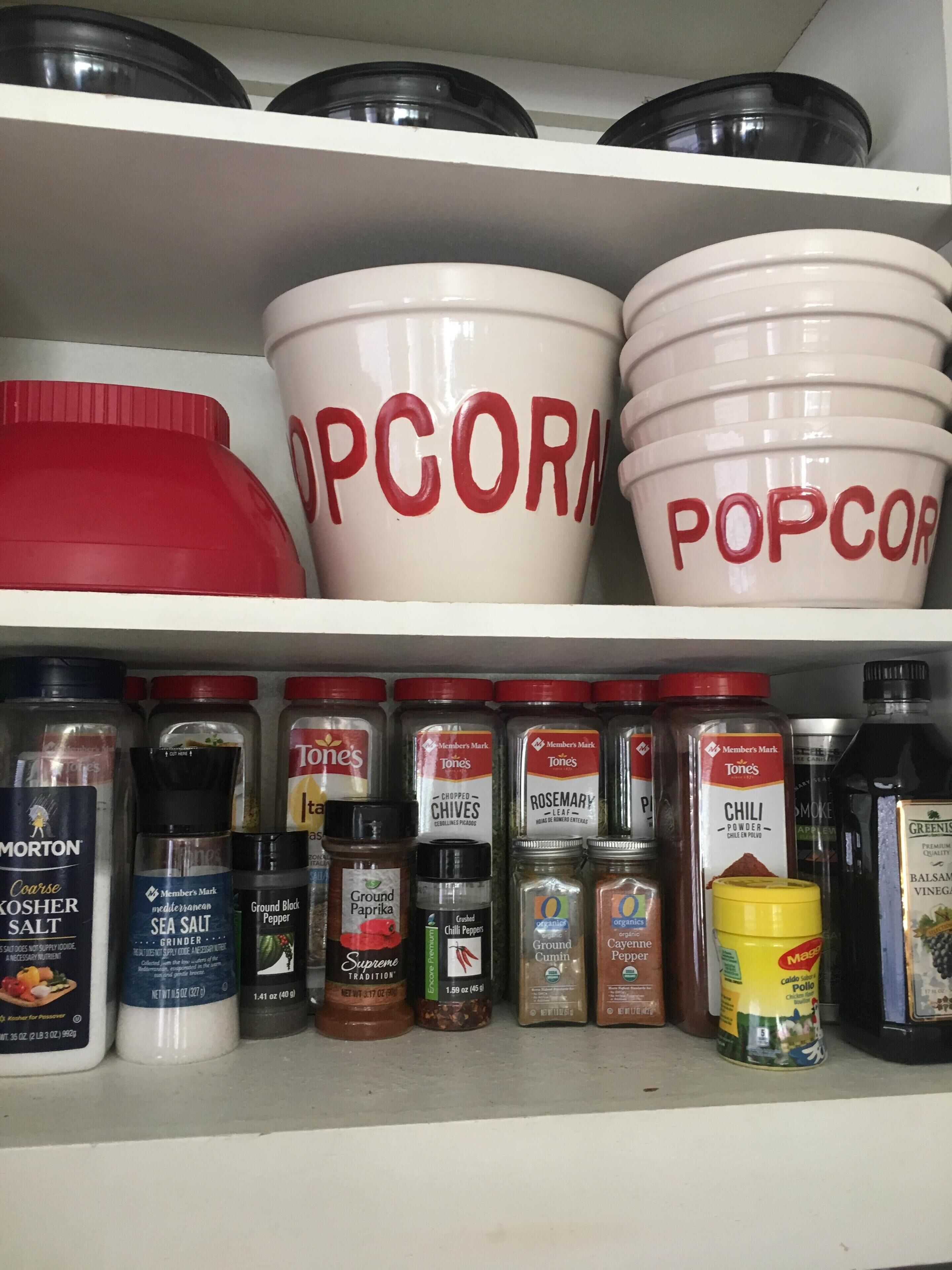 Una estantería con diversos condimentos y recipientes de cocina, incluyendo grandes tazones blancos con la etiqueta 'POPCORN' y pequeños frascos de especias como 'Pimienta Negra Molida' y 'Chile en Polvo'.