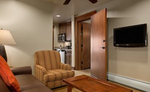 Una sala de estar con un sofá, un sillón a rayas, una mesa de centro de madera, un televisor montado en la pared y una cocina visible a través de una puerta abierta.