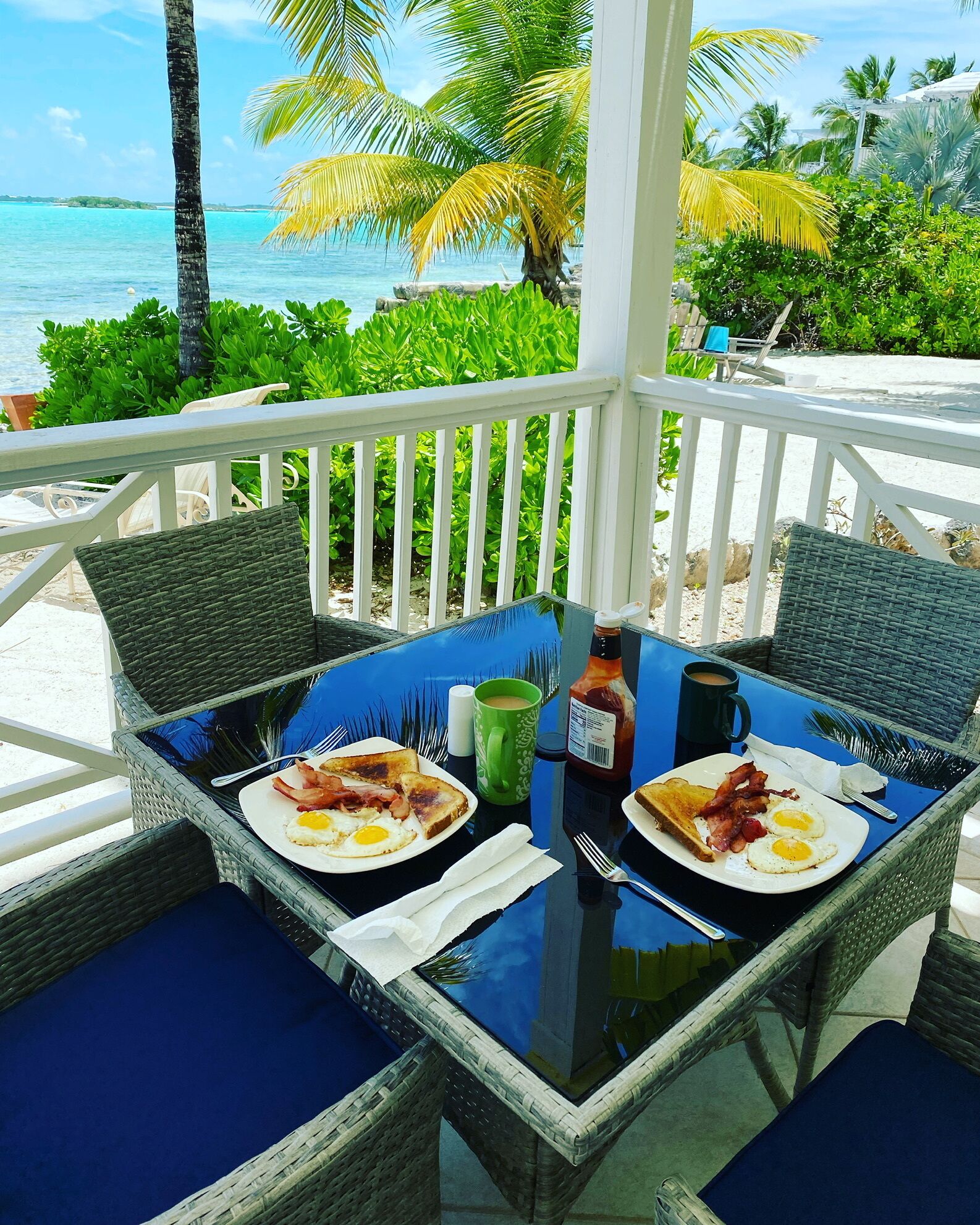 A glass-top table with wicker chairs and blue cushions, set on a balcony overlooking a beach with palm trees and the ocean.
