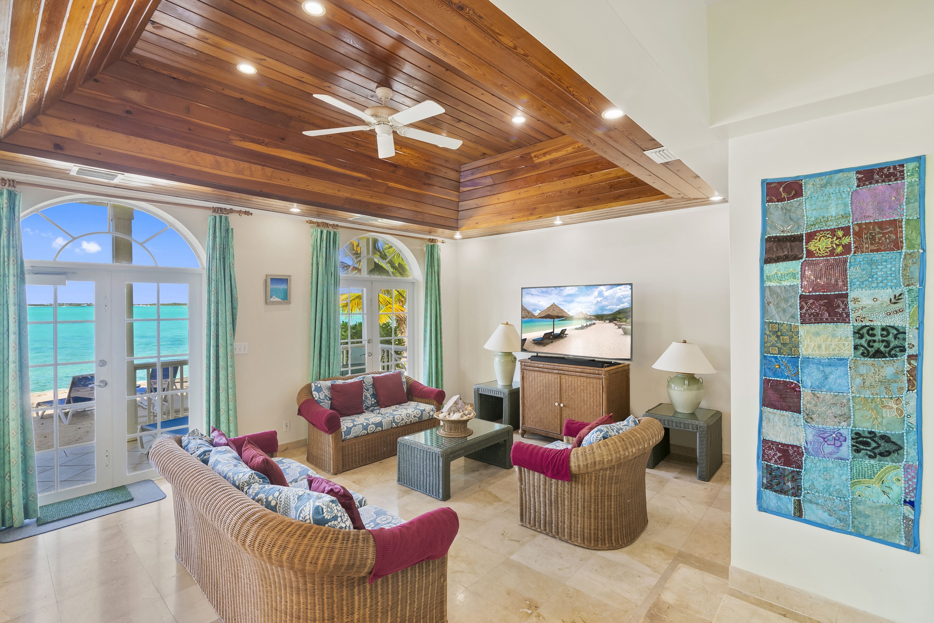A spacious living area with a wooden ceiling, wicker furniture, a television, and a large window overlooking the sea.