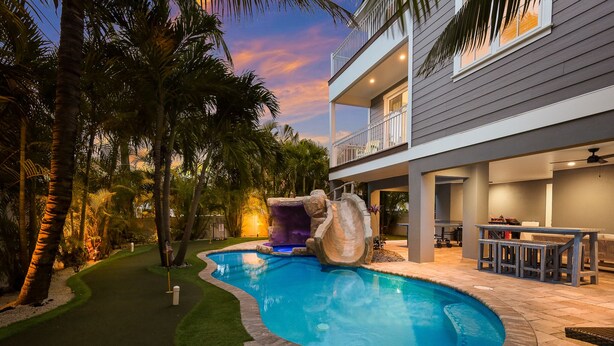 A swimming pool with a waterfall feature, surrounded by palm trees and a well-lit patio area.
