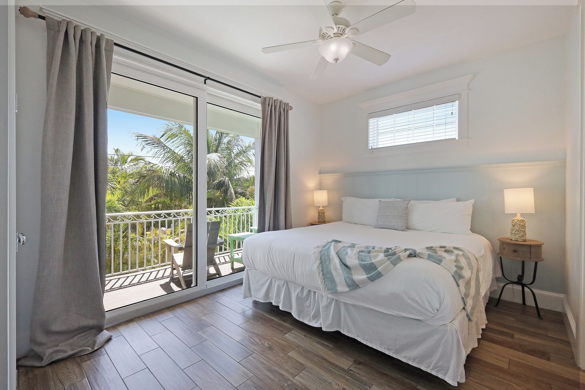 Une chambre à coucher comprenant un lit, une table de chevet, un ventilateur de plafond, une fenêtre et un balcon avec vue sur les palmiers.