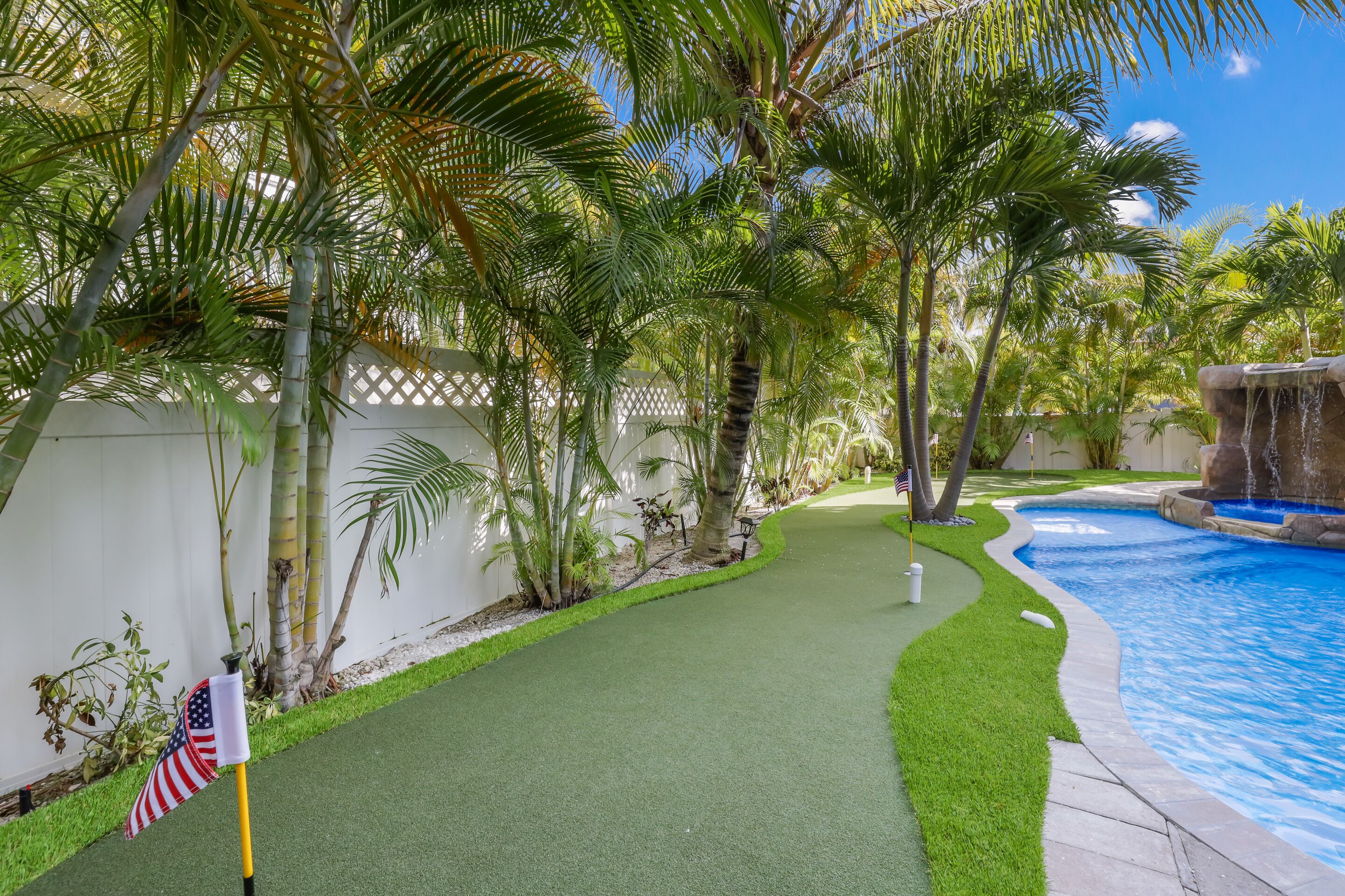 A backyard with a swimming pool, artificial grass, palm trees, and a white fence.