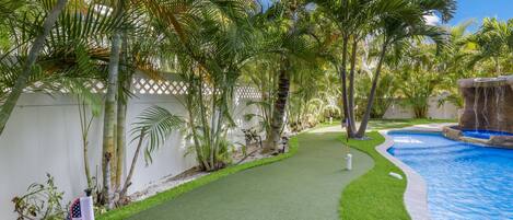 A backyard with a swimming pool, artificial grass, palm trees, and a white fence.