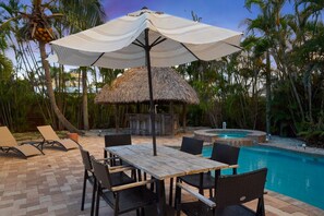 Un área junto a la piscina con mesa y sillas de madera, un parasol de paja y una piscina.