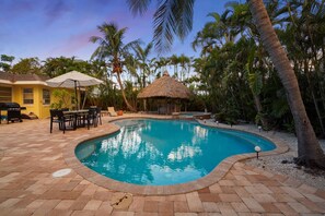 Una piscina con una cabaña de techo de paja, rodeada de palmeras y un patio con sillas y una mesa.