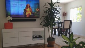 A living room with a flat-screen TV displaying a show, a white entertainment unit, a potted plant, and a dining area with chairs and a table.