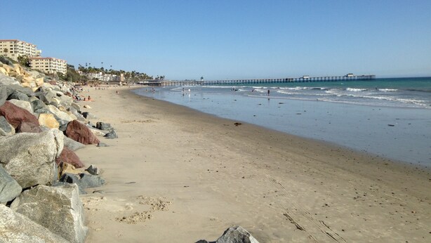 A beach with a rocky shoreline, a pier extending into the water, and buildings in the background.