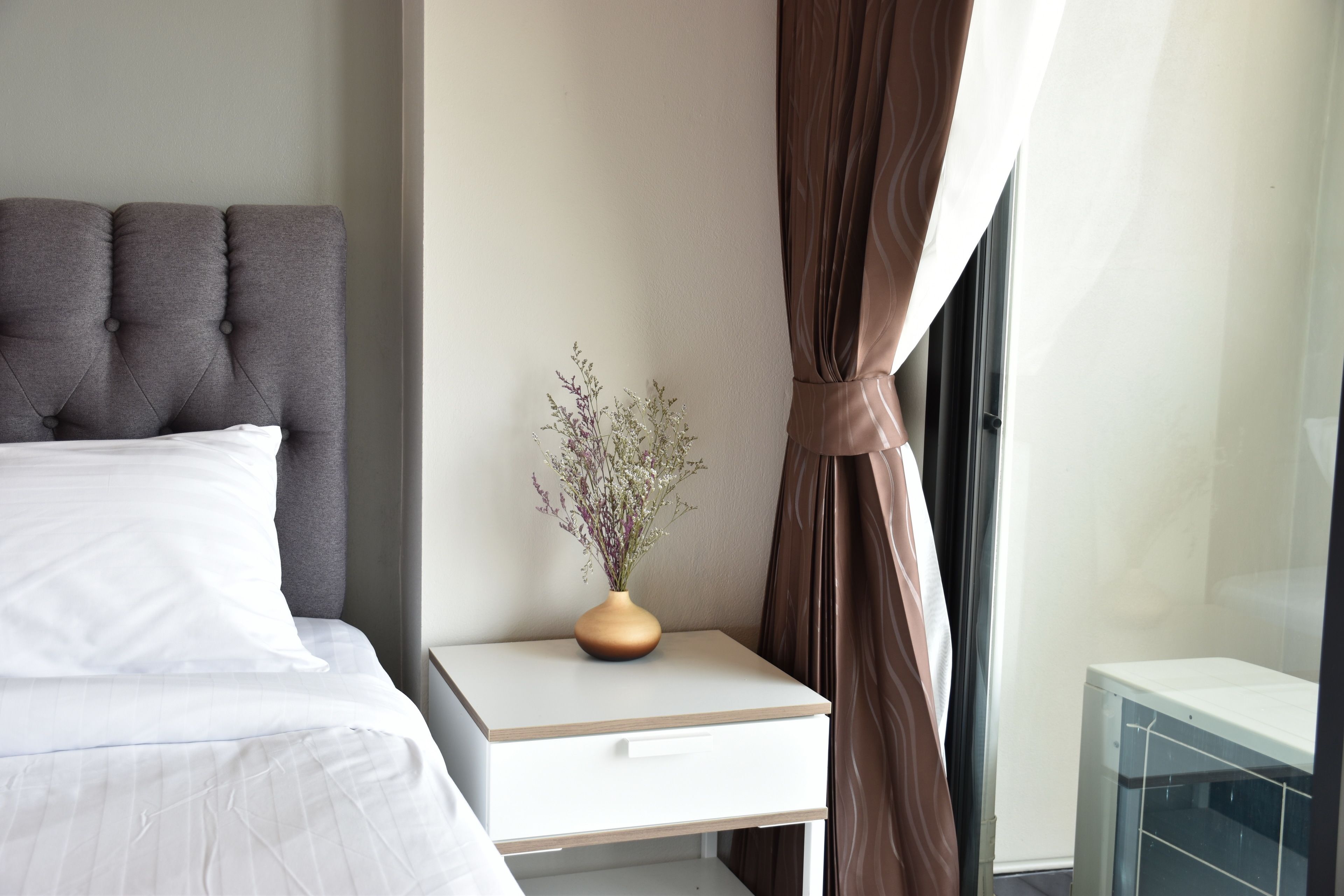 Una cama bien hecha con cabecera gris, una mesita de noche con un florero y una ventana con cortinas marrones.