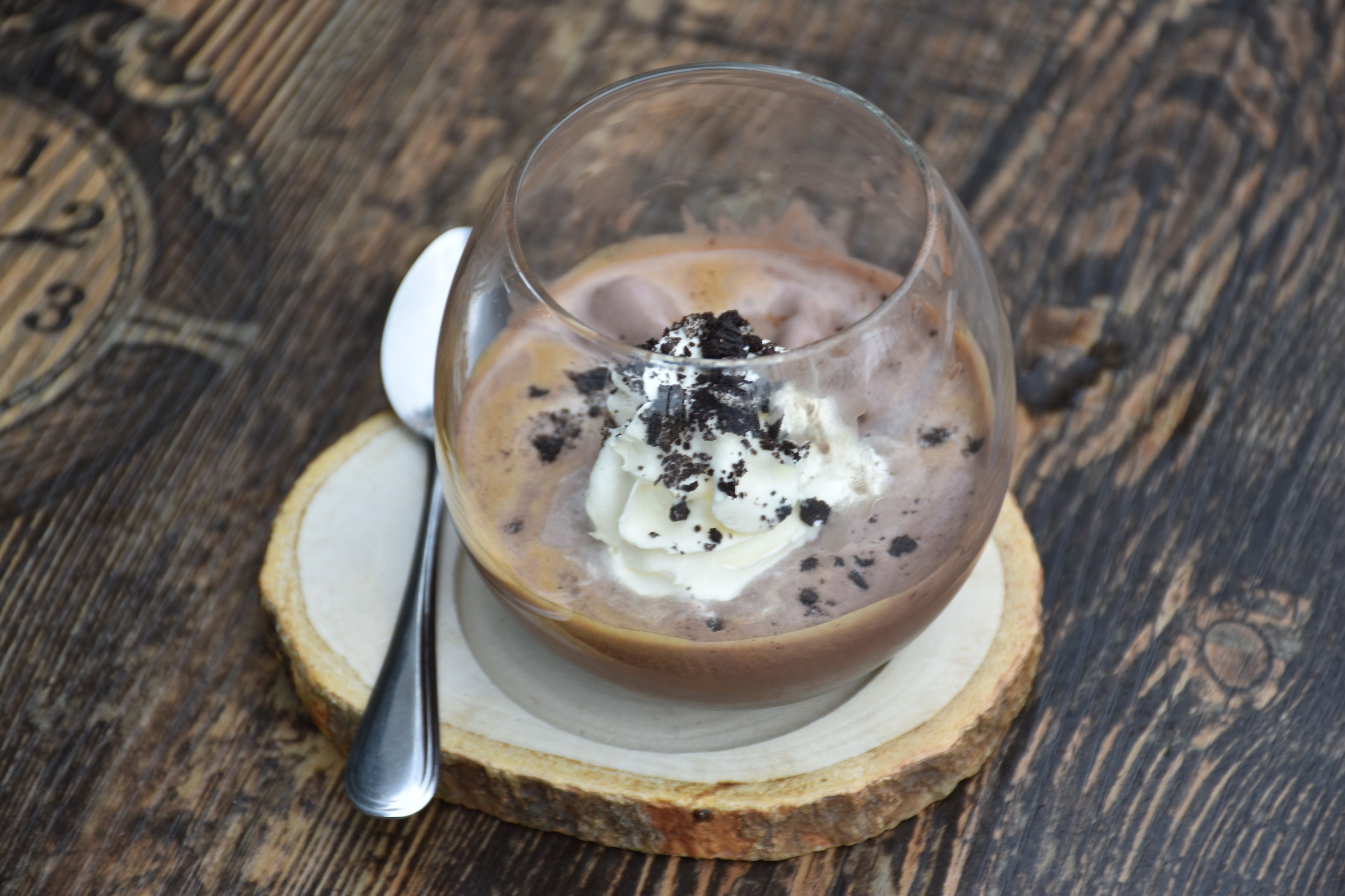 A dessert in a glass on a wooden coaster with a spoon beside it.