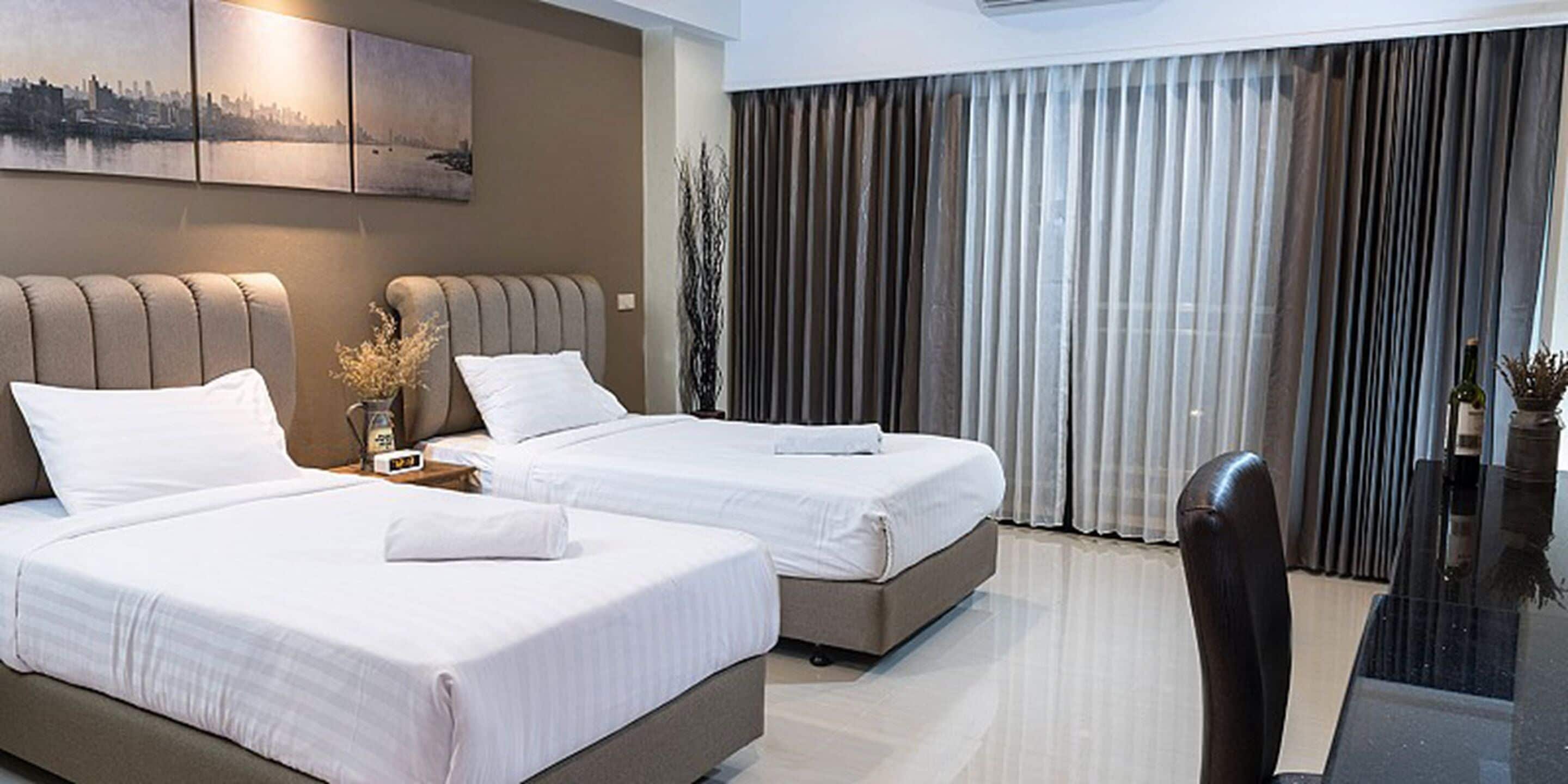 A hotel room with two beds, a large window with curtains, a desk with a chair, and a cityscape painting on the wall.