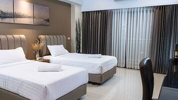 A hotel room with two beds, a large window with curtains, a desk with a chair, and a cityscape painting on the wall.