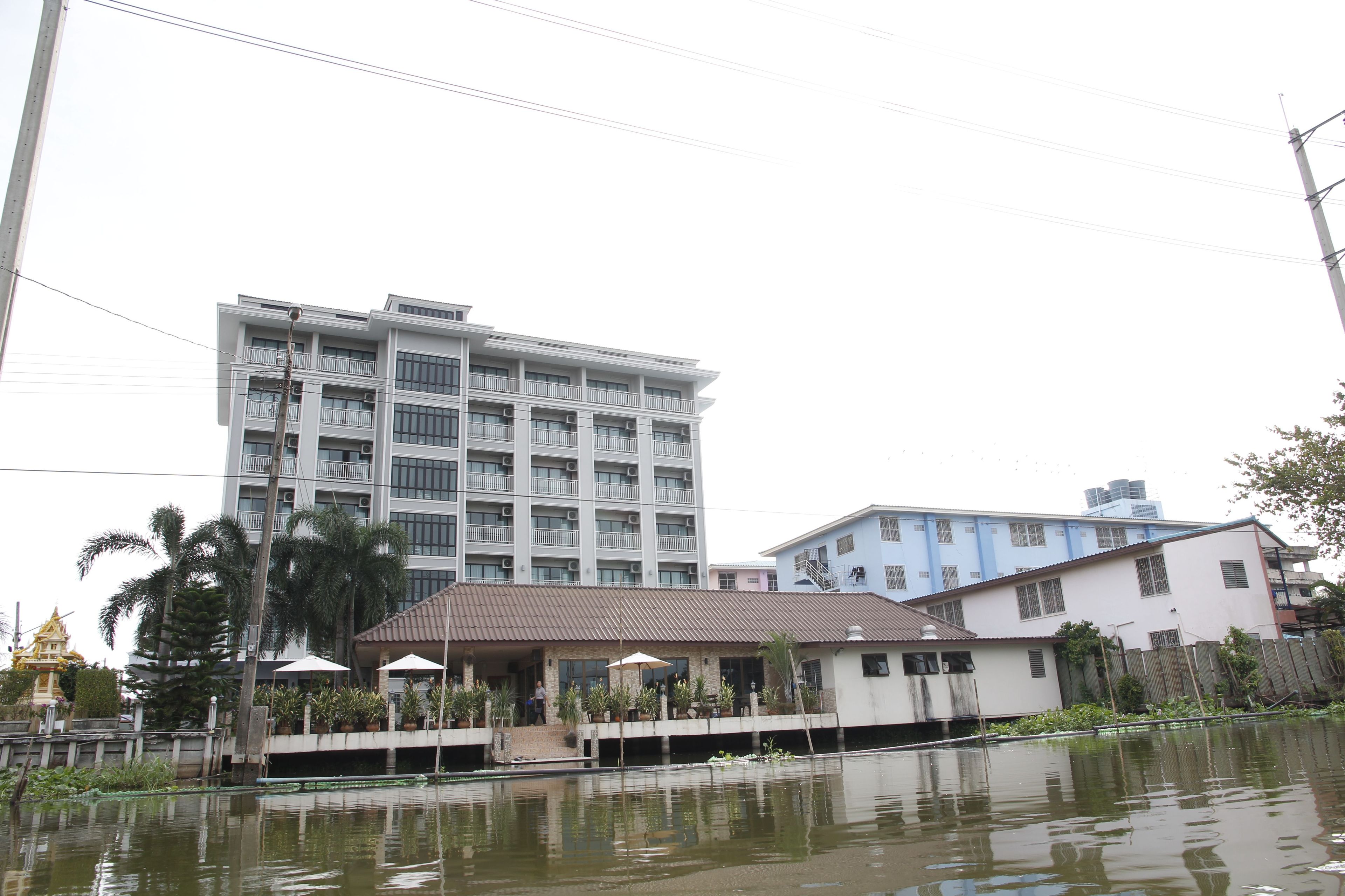 A multi-story hotel with balconies, a covered outdoor area with umbrellas, and a body of water in front.