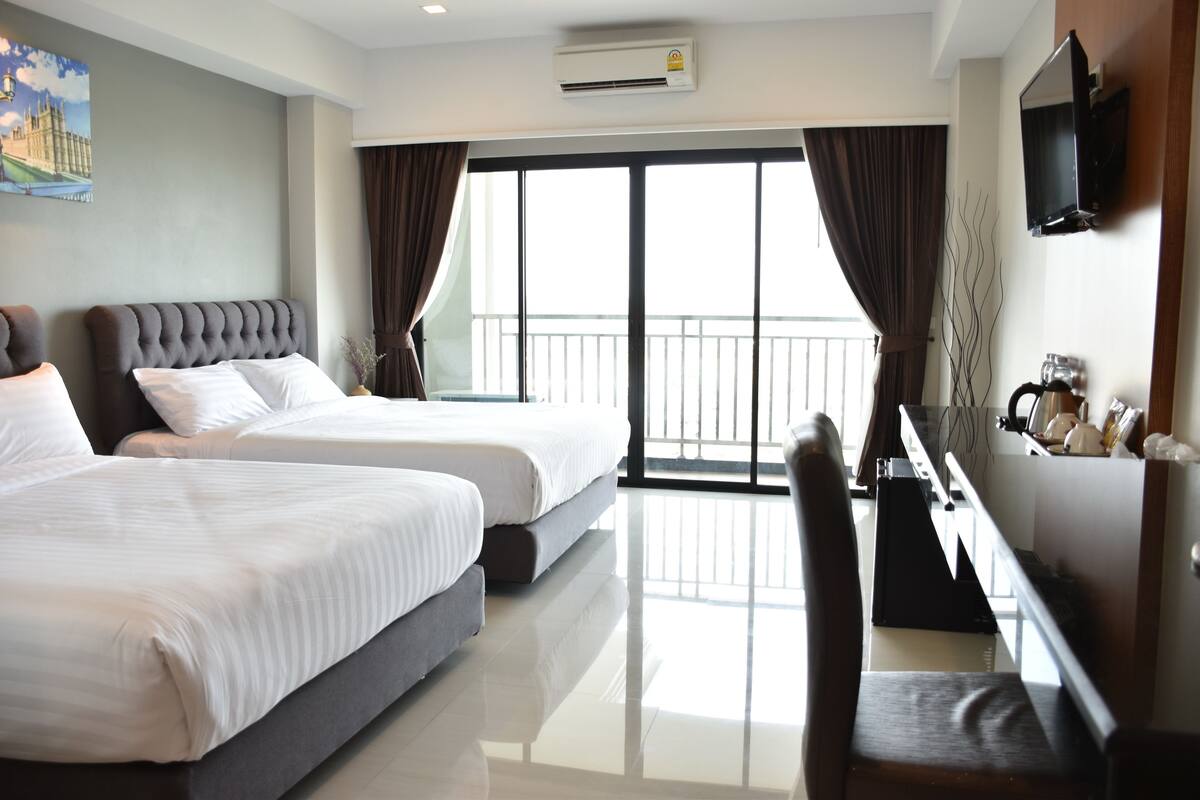 A hotel room with two beds, a TV, a desk, and a balcony with curtains.