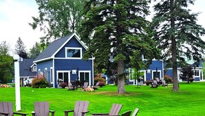 Een blauw gebouw met een schuin dak en een terras met Adirondack-stoelen en een vuurkuil.